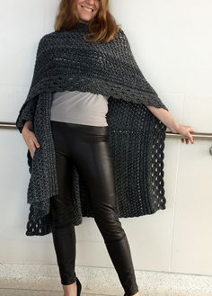 a woman standing in front of a white wall wearing black leather pants and a knitted shawl