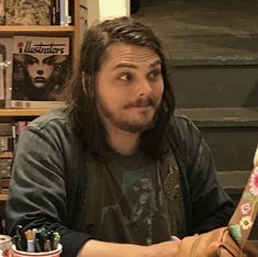 a man with long hair sitting at a table in front of a bookshelf