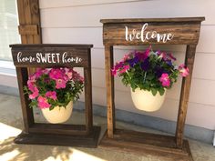 two flower pots with flowers in them sitting next to a welcome sign on the side of a house