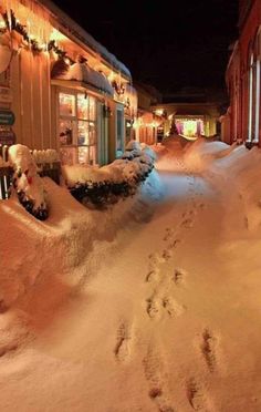 a snow covered street with footprints in the snow and buildings lit up by christmas lights