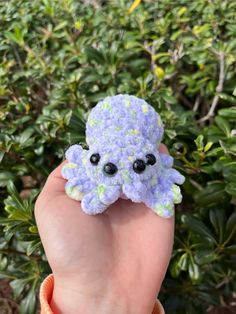 a hand holding a small blue and green crocheted octopus brooch on it's finger