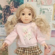 a doll with blonde hair wearing a pink sweater and plaid skirt, standing in front of a wall full of pictures