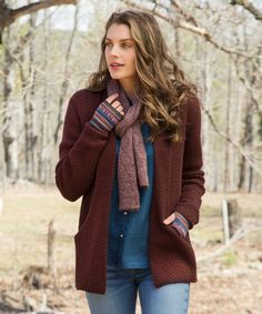 a woman standing in the woods wearing a sweater and scarf with mittens on her hands