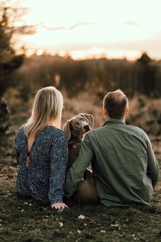 a man and woman sitting on the ground with a dog in their lap looking at the sunset