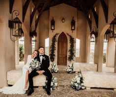 a newly married couple sitting on a bench in front of a building with flowers and greenery