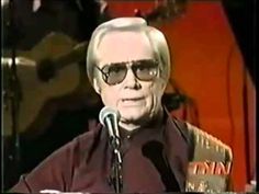 an older man wearing sunglasses and holding a microphone in front of a microphone with a guitar behind him