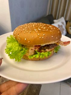 a hamburger with lettuce, bacon and cheese on a white plate in someone's hand