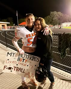 two people posing for a photo with a football sign in front of a fence at night