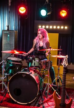 a woman with pink hair playing the drums in front of a microphone and some lights