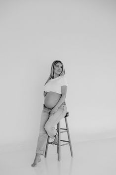 a pregnant woman sitting on top of a stool in front of a white wall and posing for the camera