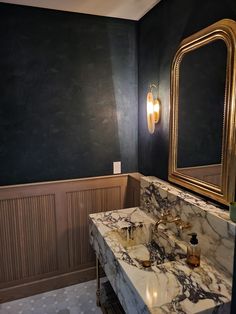 a bathroom with marble counter top and gold framed mirror on the wall, along with black walls