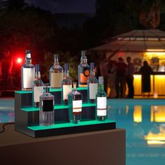 several bottles and glasses sitting on top of a shelf next to a pool at night