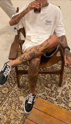 a man with tattoos sitting in a chair drinking from a beer glass while holding a cell phone to his ear