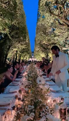 two men in tuxedos standing at the end of a long table with food on it