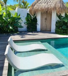 two chaise lounges sitting next to a swimming pool in front of a thatched hut