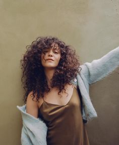 a woman with long curly hair wearing a brown dress and blue jacket leaning against a wall