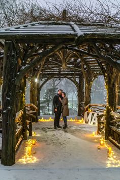 two people standing under a wooden gazebo covered in snow and lite - up lights