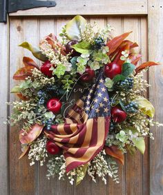 a wreath with an american flag and apples hanging on a wooden door, surrounded by foliage