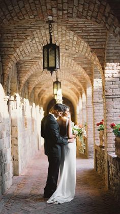 a bride and groom are standing in an archway between two brick buildings with lanterns hanging from the ceiling