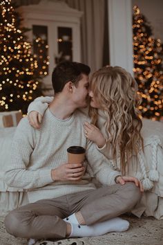 a man and woman sitting on the floor kissing while holding a coffee cup in front of christmas trees