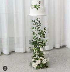 a three tiered cake with greenery and white flowers on the top is sitting in front of a window