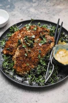 a black plate topped with meat covered in greens and sauces next to a bowl of mustard