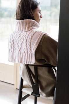 a woman sitting in a chair wearing a pink cabled sweater and looking out the window