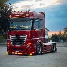 a red semi truck parked in a parking lot next to a fence and trees at sunset