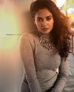 a woman with long hair wearing a gray sweater and silver necklace, leaning against a wall