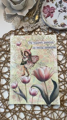 a card with a fairy on it sitting next to some plates and flowers in the background