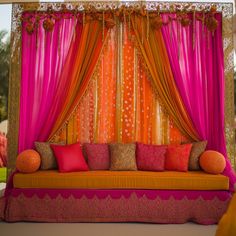 an orange and pink couch sitting under a window covered in drapes with pillows on it