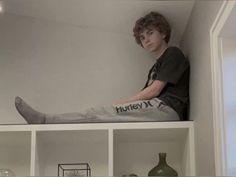 a young man sitting on top of a book shelf next to a vase and other items