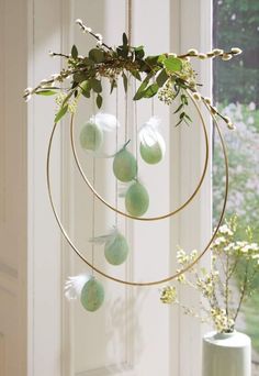 a white vase sitting next to a window filled with flowers and green eggs hanging from a gold hoop