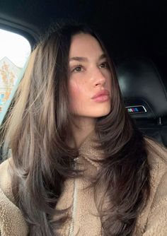 a woman sitting in the back seat of a car with long dark hair and wearing a beige coat