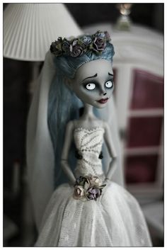 a doll with blue hair wearing a white dress and flowers in her hair is standing next to a lamp