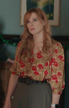 a woman with long red hair wearing a floral shirt and brown skirt standing in a living room