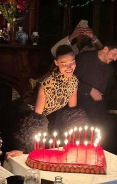 a woman sitting in front of a red cake with lit candles on it, surrounded by other people