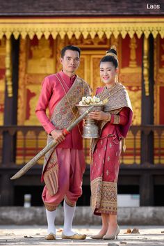 a man and woman dressed in traditional thai garb holding a silver bowl with flowers on it