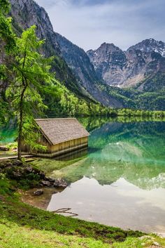 a small cabin sitting on the shore of a lake surrounded by mountains and greenery