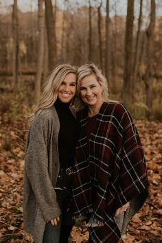 two women standing next to each other in the woods with fall leaves on the ground