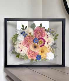 an arrangement of flowers in a black frame on a wooden table next to a mirror