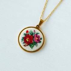 a cross stitch necklace with flowers in the center on a gold plated chain hanging from a white surface