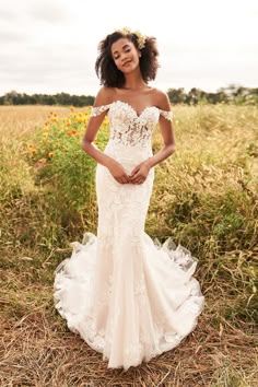 a woman standing in a field wearing a wedding dress with an off the shoulder design