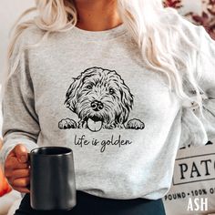 a woman holding a black coffee mug in her right hand and wearing a grey sweatshirt with the words life is golden on it