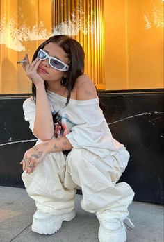 a woman sitting on the ground with her hand to her face, wearing white pants and sunglasses