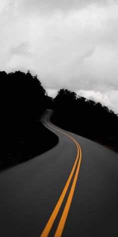 an empty road with two yellow lines in the middle and trees on both sides, against a cloudy sky