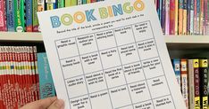 a person holding up a book bingo sheet in front of a bookshelf full of books