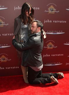 a man kneeling down on the red carpet with his arm around a woman's shoulder