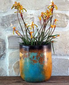 an orange and blue vase filled with flowers on top of a wooden table next to a stone wall