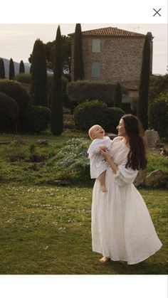 a woman in a white dress holding a baby up to her chest and looking at the sky